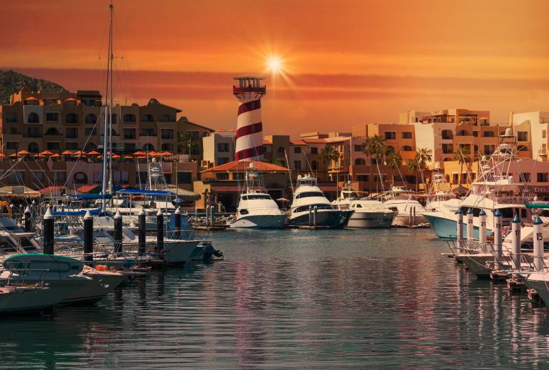 A vibrant sunset casts a warm glow over a picturesque harbor filled with boats and a striking lighthouse.