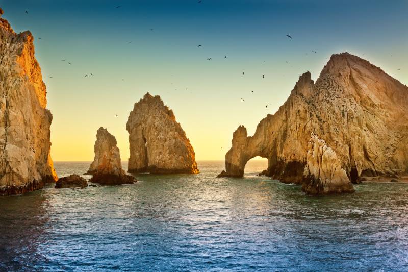 Golden hour light illuminates dramatic rock formations and the iconic arch at Land's End in Cabo San Lucas.