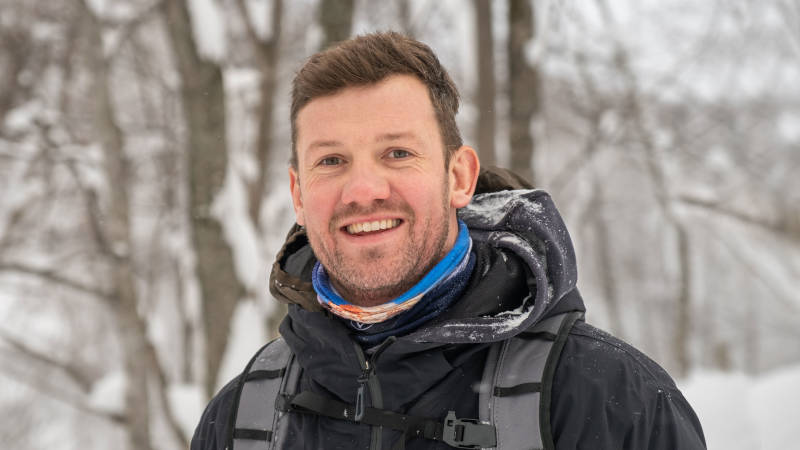 A smiling man bundled in a winter jacket and backpack poses for a photo amidst a snowy forest.