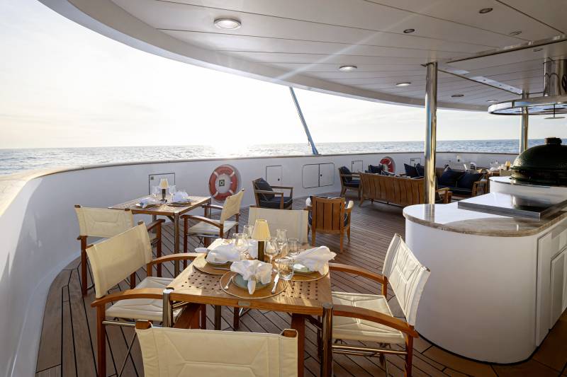 A cozy outdoor seating area on the deck of a boat, with wooden furniture, a bar, and a scenic view of the water in the background.