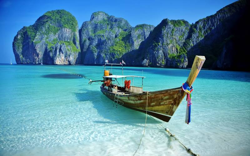 A traditional longtail boat rests in the crystal-clear turquoise waters of a tropical bay, with towering limestone cliffs rising majestically in the background.