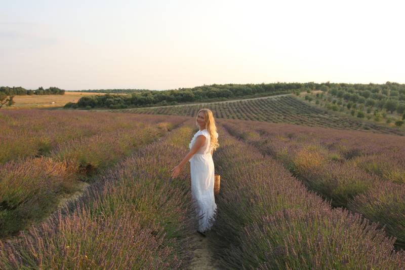A woman in a white dress stands in a lavender field with rows of purple flowers stretching into the distance under a hazy sky.