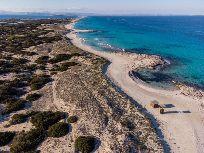 Sandy dunes dotted with scrub brush lead to a pristine white beach and turquoise waters, with a distant coastline under a clear blue sky.