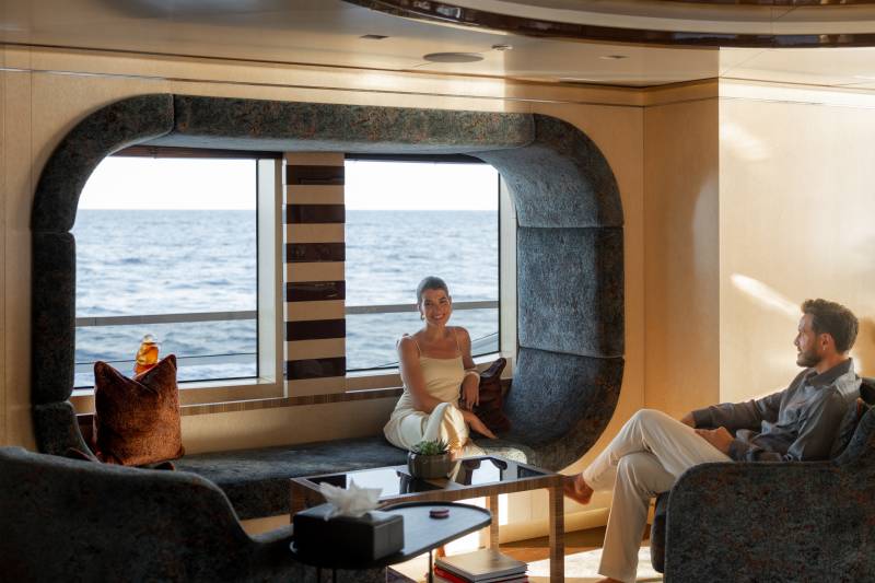 A couple relaxes in a luxurious yacht lounge with a large window overlooking the ocean.
