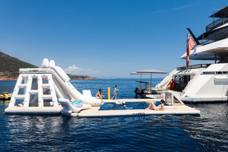 People relax on an inflatable water park and pool attached to a large yacht, with a hilly coastline in the background.