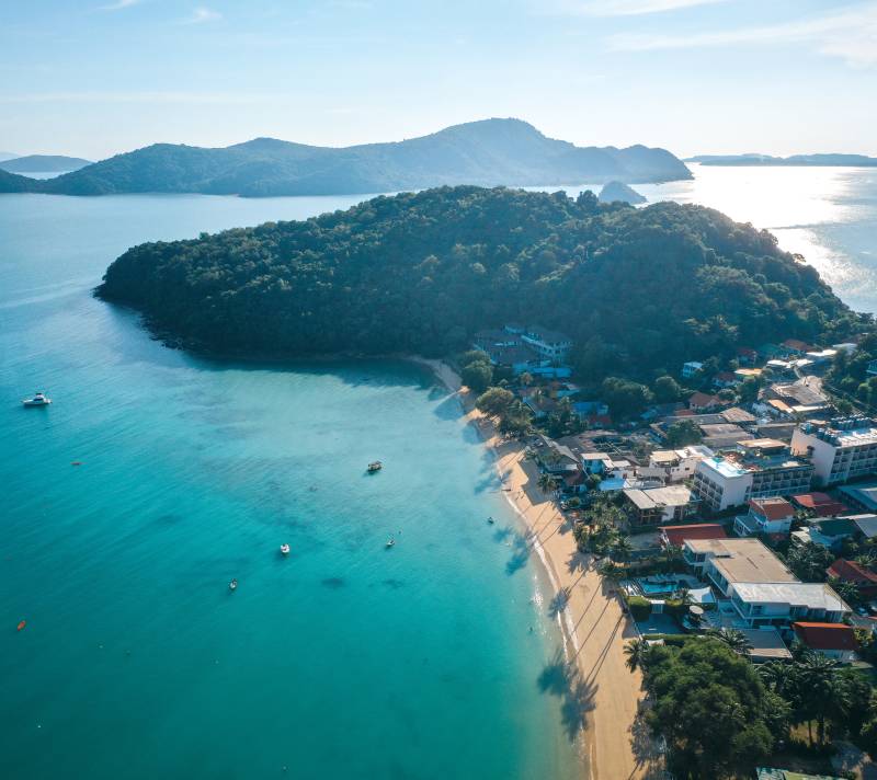 A crescent-shaped sandy beach with buildings and palm trees lines the turquoise ocean, with lush green islands in the distance under a clear blue sky.