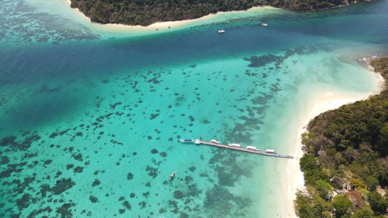 A long pier with several boats extends into the crystal-clear turquoise water, with lush green islands forming the background.