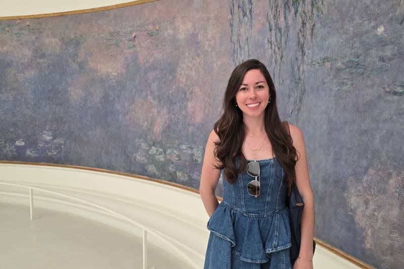 A smiling woman in a denim dress stands in front of a large, impressionistic painting of water lilies.