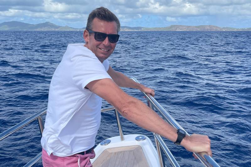 A man in a white t-shirt and pink shorts smiles while leaning on a railing on a boat, with a blue ocean and distant islands in the background.