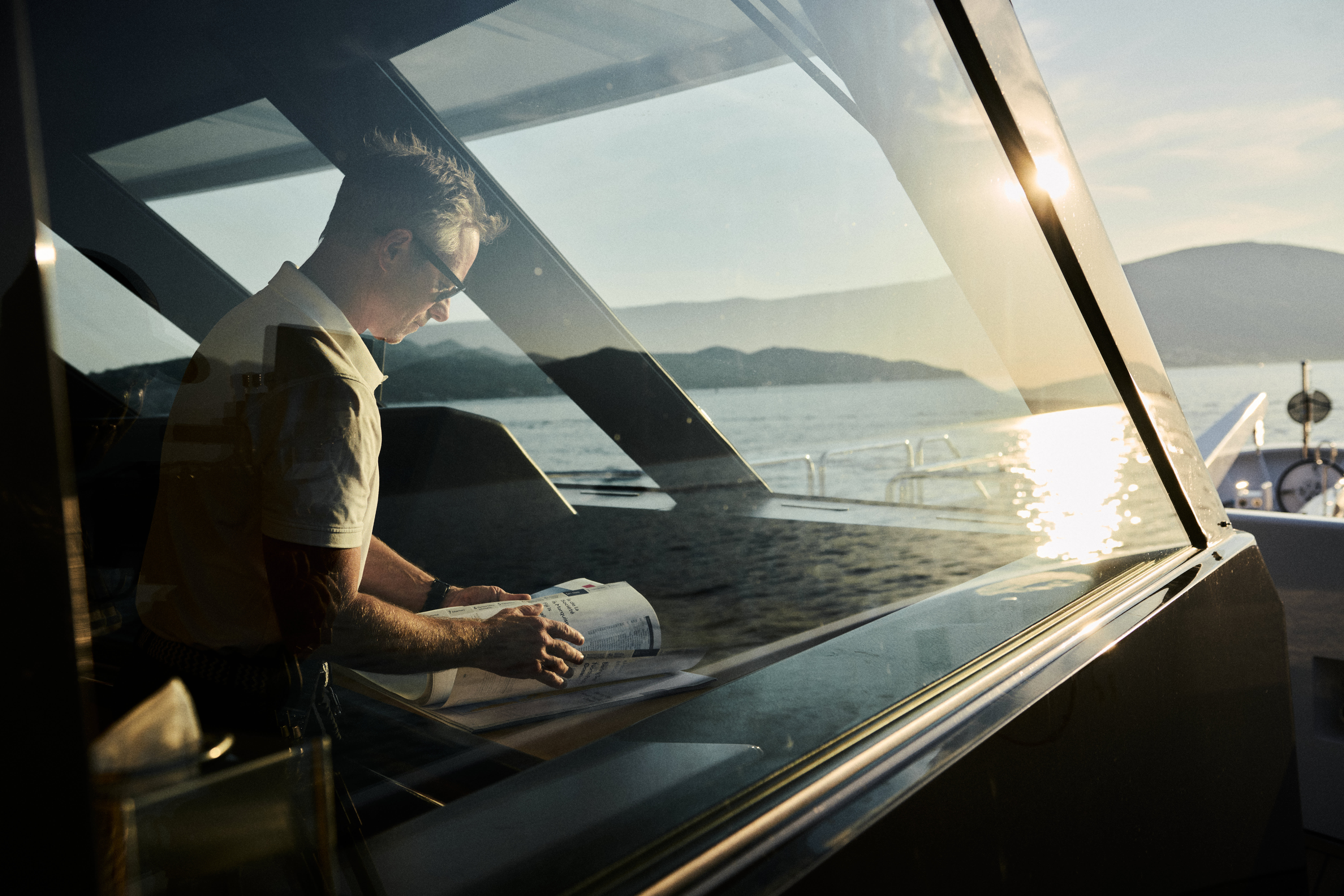 A person is sitting by a window, gazing out at a scenic landscape with mountains and water in the background, as the sun sets in the distance.